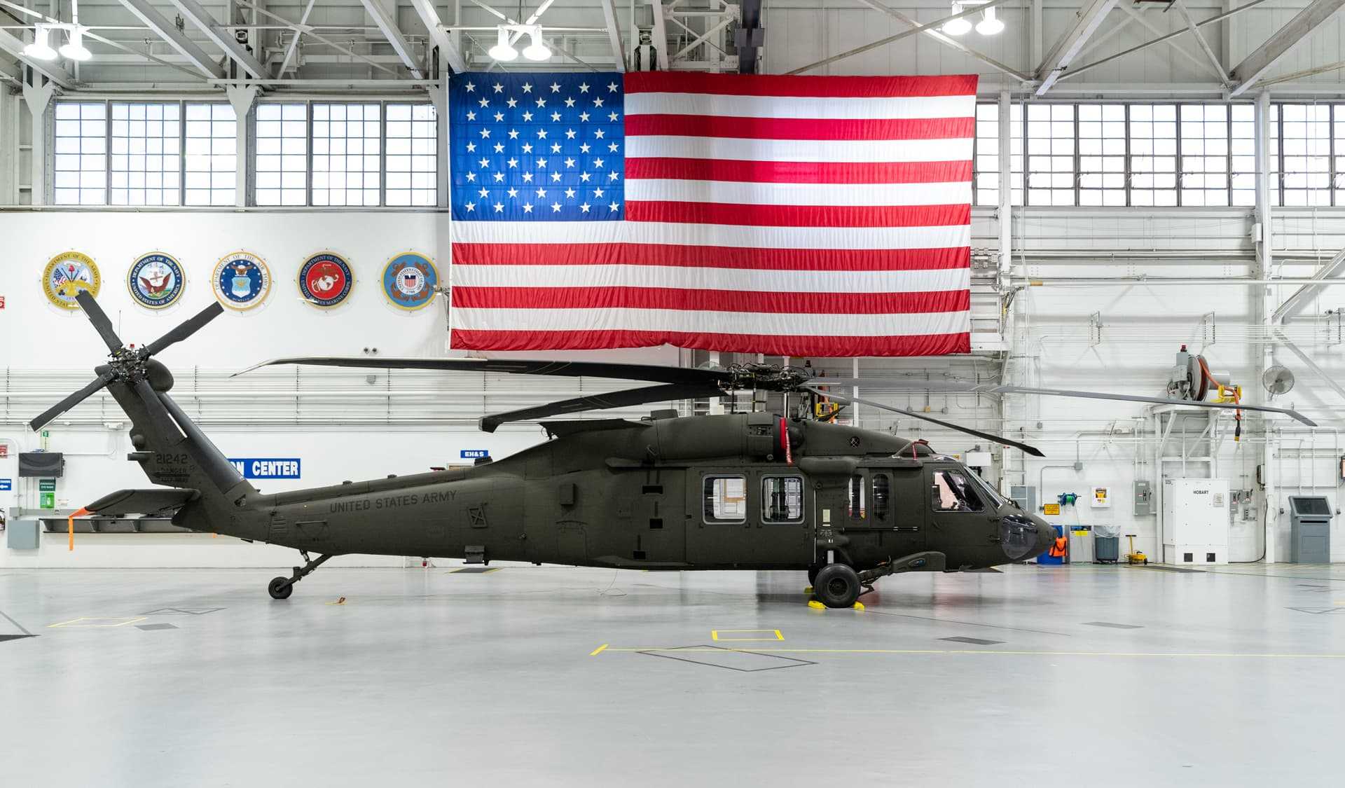 A Black Hawk UH60 helicopter inside a hangar, a big flag of the United States of America is in the wall located behind the helicopter